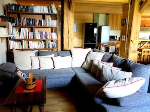 Una sala de estar acogedora con un sofá modular, una estantería de madera llena de libros y una zona de cocina al fondo.