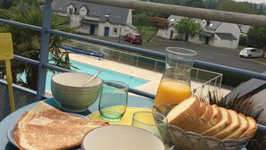 Une table de petit-déjeuner avec des toasts, un bol, un verre et une carafe de jus, le tout placé sur un balcon donnant sur une piscine et des maisons.