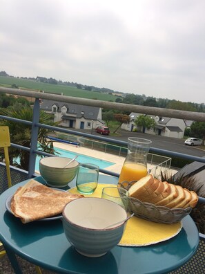 Et morgenbord med ristet brød, en skål, et glas og et juice kander på en balkon med udsigt over en swimmingpool og huse.