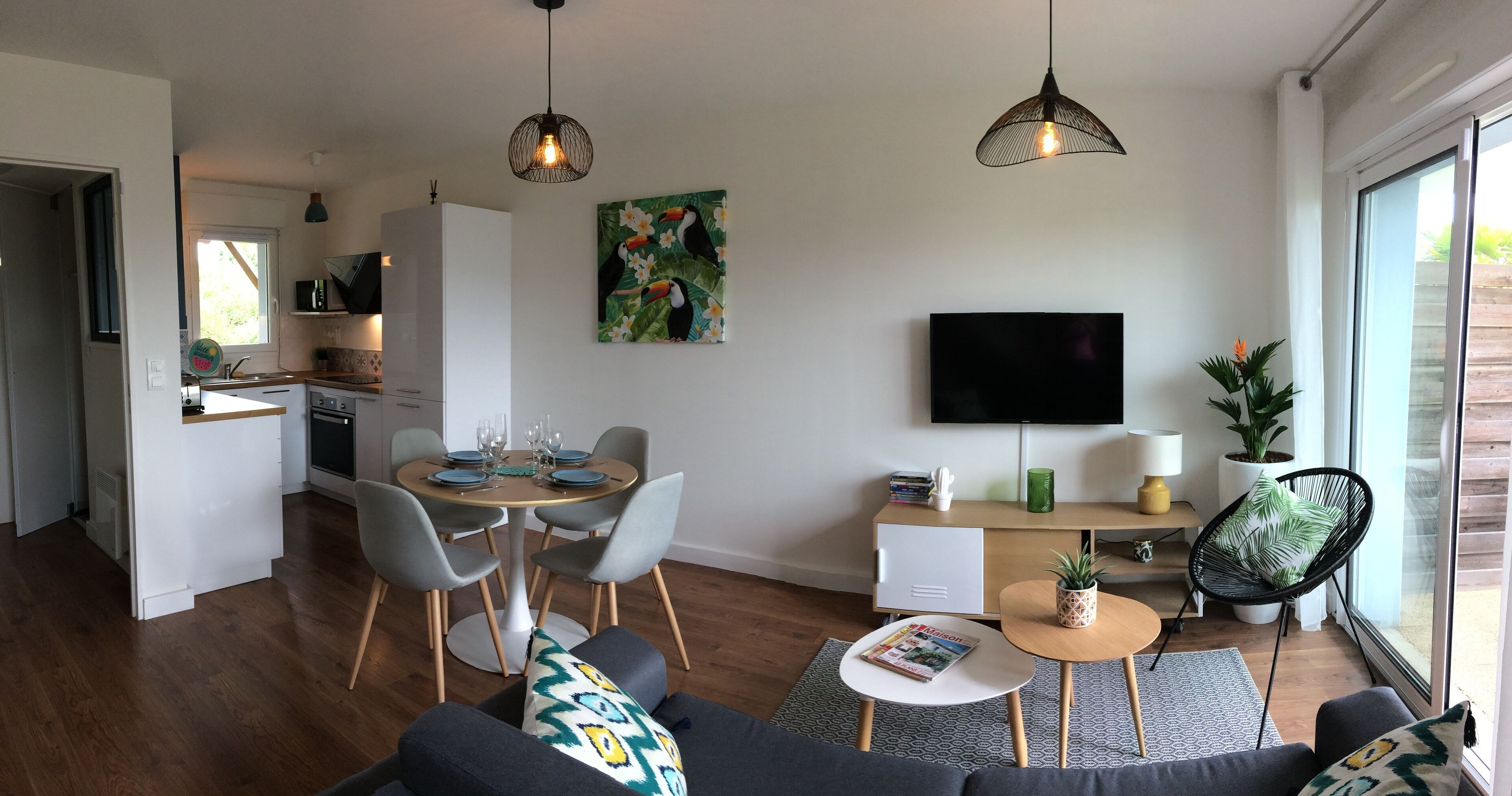 Ein modernes Wohnzimmer mit einer Couch, einem Couchtisch und einem Essbereich mit Stühlen und einem Tisch für vier Personen. An der Wand ist ein Fernseher befestigt und auf einem Beistelltisch steht eine Zimmerpflanze.