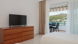 A hotel room with a TV on a wooden dresser, a bed, a balcony with a table and chairs, and a view of a hillside. - Villa with private swim pool, 40 meters from the sea (Seget Vranjica)