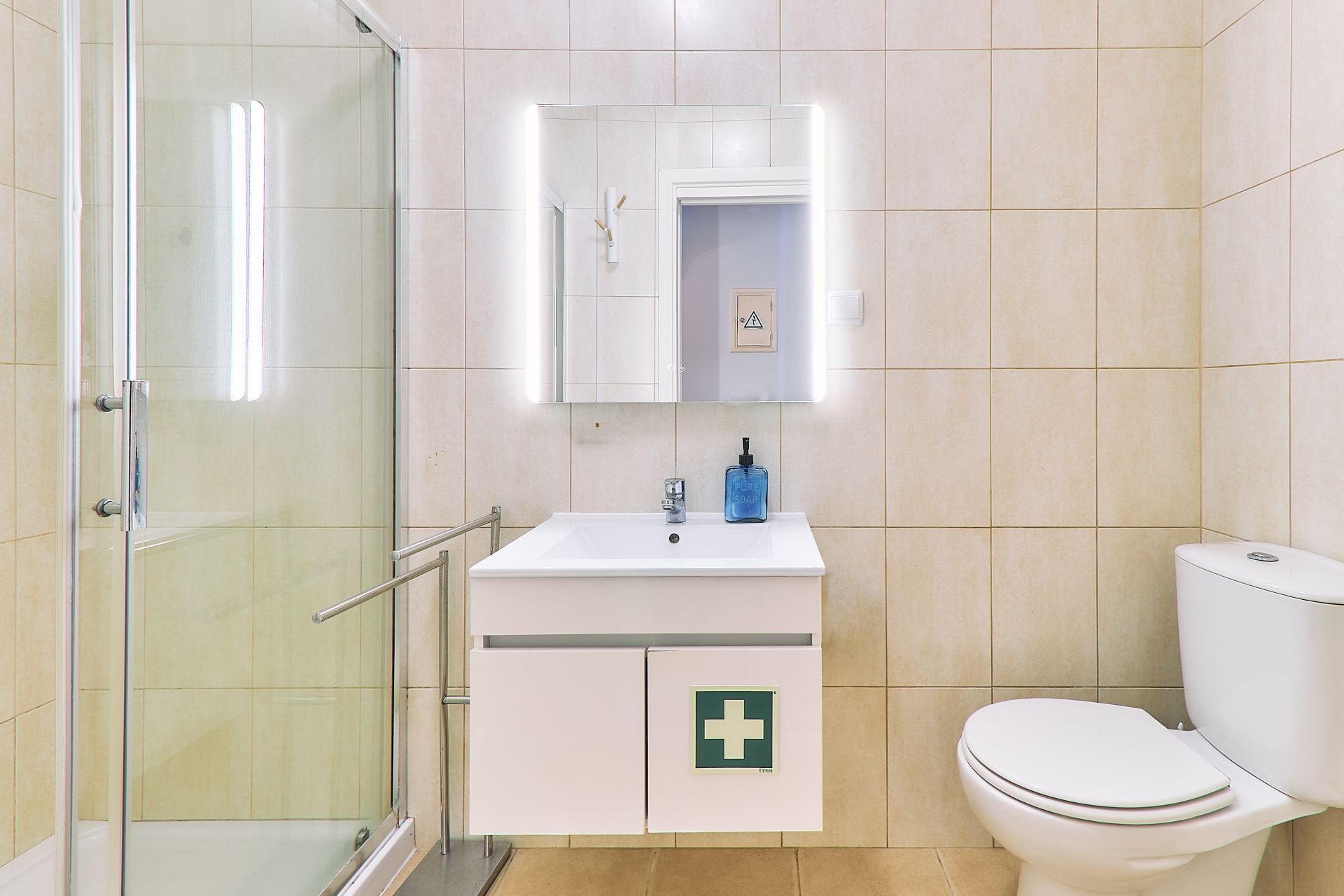 Ein Badezimmer mit Duschkabine aus Glas, einem weißen Waschbecken mit Seifenspender und einer Toilette mit grünem Notfallschild.
