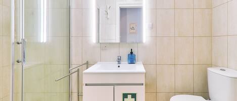 Ein Badezimmer mit Duschkabine aus Glas, einem weißen Waschbecken mit Seifenspender und einer Toilette mit grünem Notfallschild.