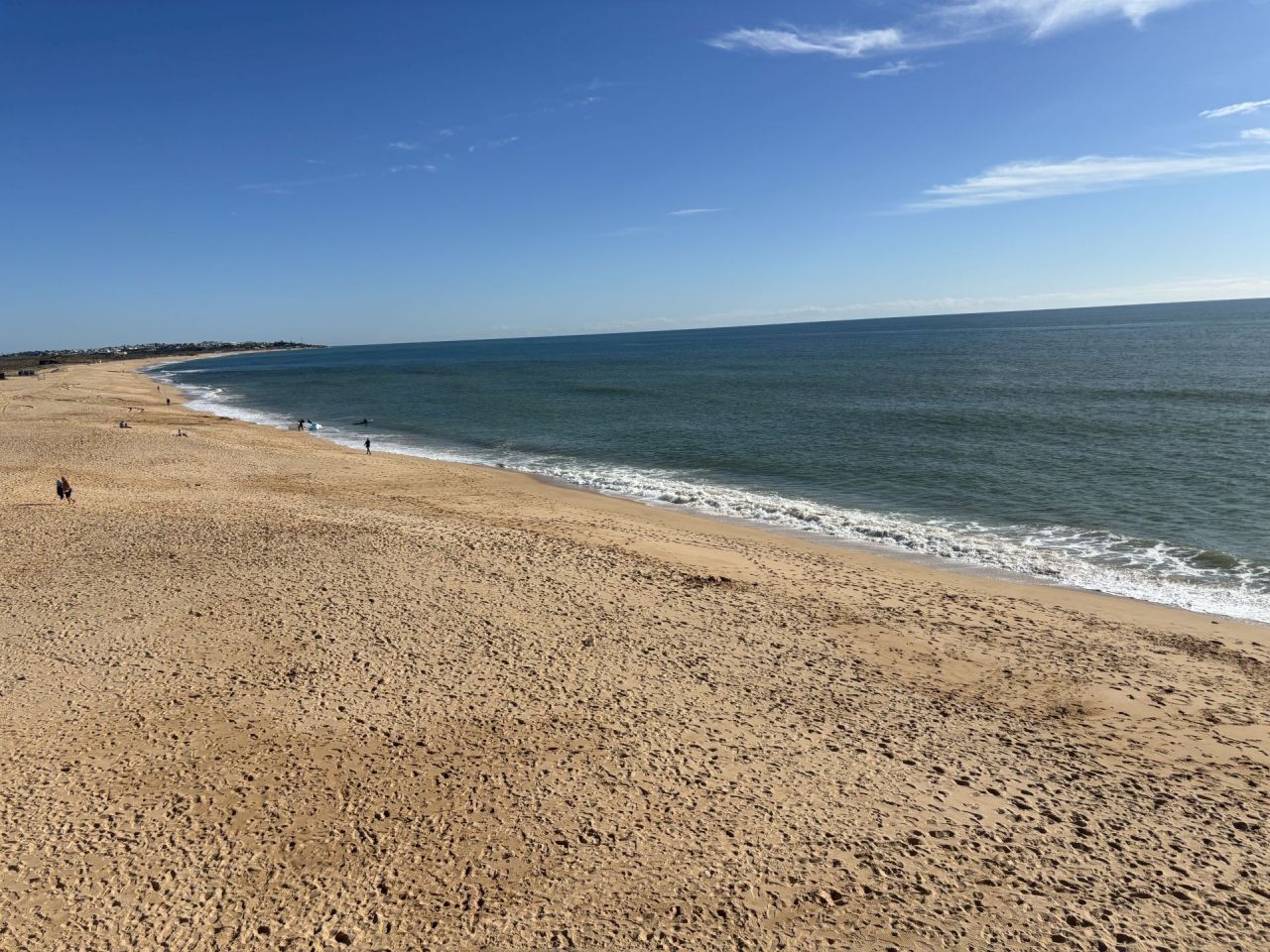 Ampia spiaggia sabbiosa con onde calme, cielo sereno e costa in lontananza.