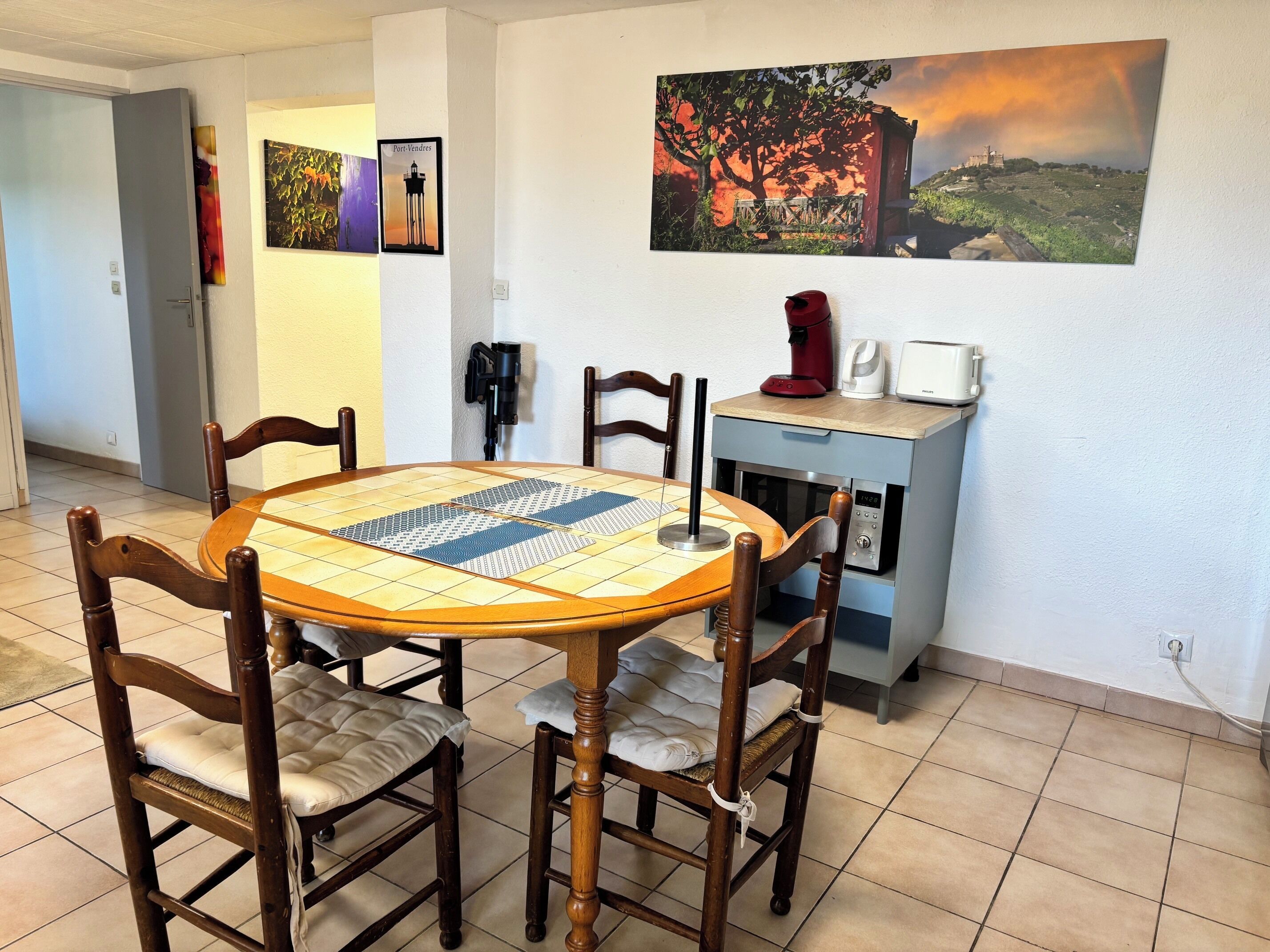 A dining area with a round table, four wooden chairs, a coffee machine, a toaster, and a painting on the wall.