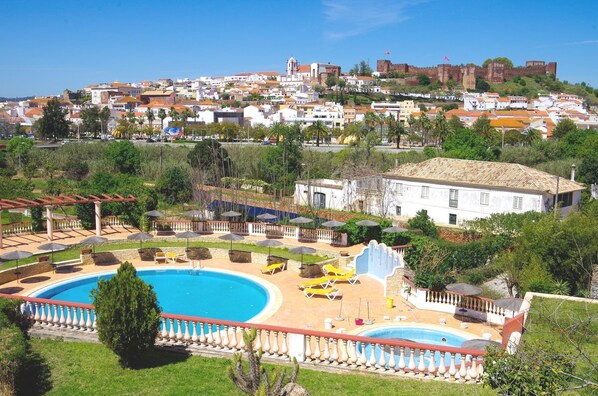 A resort with a large swimming pool, sun loungers, and a view of a historic town with a castle in the background.