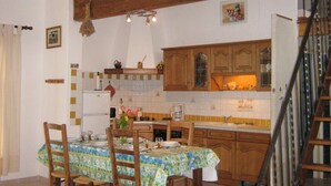 A dining area with a table set for four, wooden chairs, and a kitchen in the background with wooden cabinets and a tiled backsplash. - Comfortable gîte in the heart of the quiet luberon countryside, ideal for families. (Bonnieux)