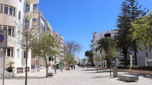 Une zone piétonne dallée, bordée de bâtiments et d’arbres, avec un ciel dégagé.