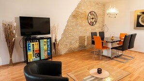 A modern living room with a flat-screen TV, a stone accent wall, a dining area with orange chairs, and a chandelier.