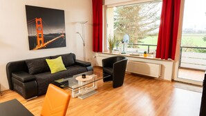 A modern living room with a black sofa, orange chairs, a glass coffee table, and a large window with red curtains.
