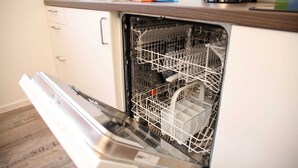 An open dishwasher with racks and utensils inside, a kitchen counter with a coffee maker, and a drawer unit.