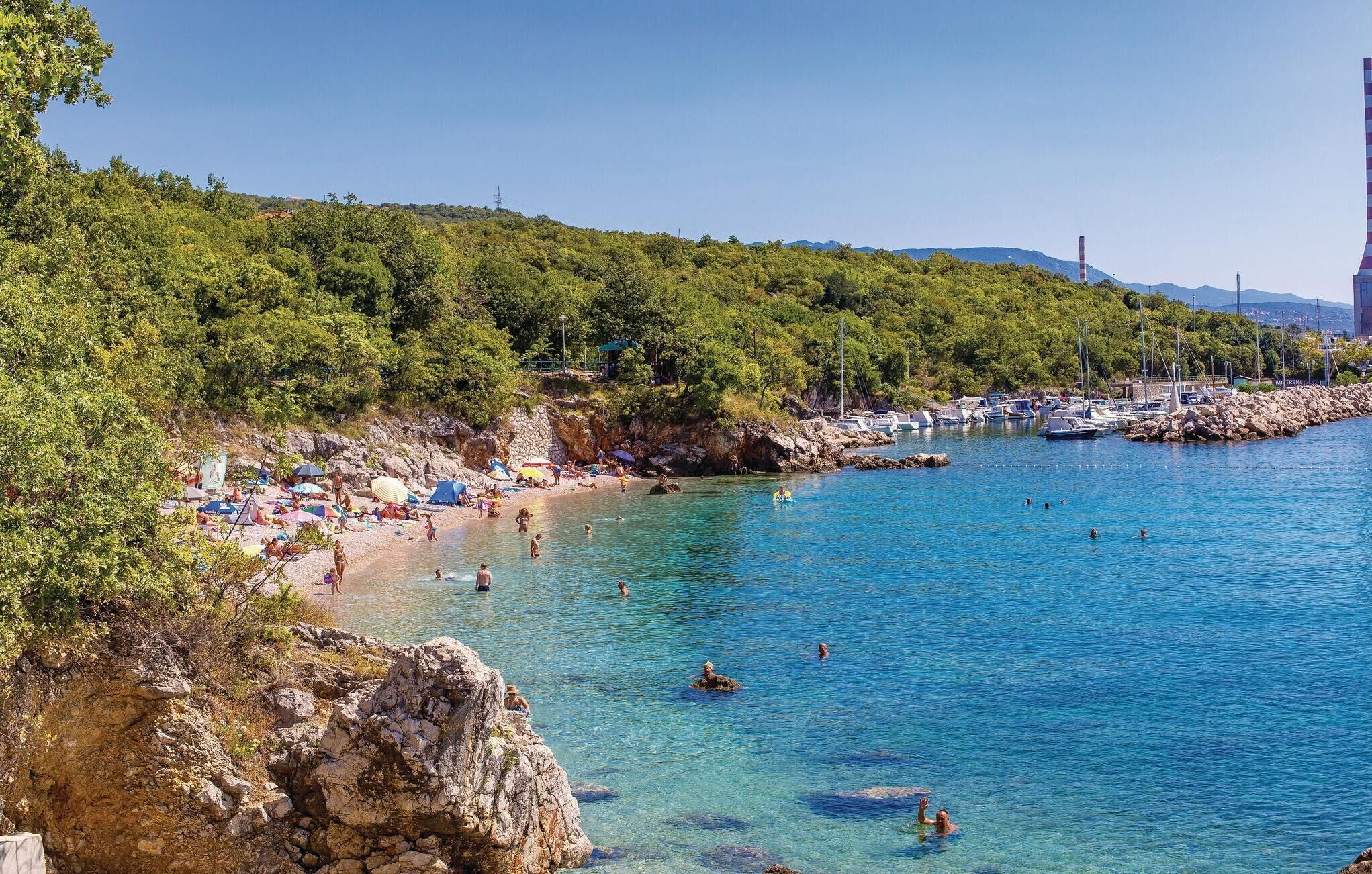 Une plage côtière bordée d’une eau bleu cristallin, d’un rivage rocheux et d’un port de plaisance où sont amarrés des bateaux.