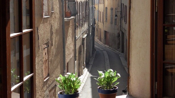 A narrow street with buildings on either side, viewed from a window with a view of two potted plants on the sill. - L'Appart, charm and confort  (Cotignac)