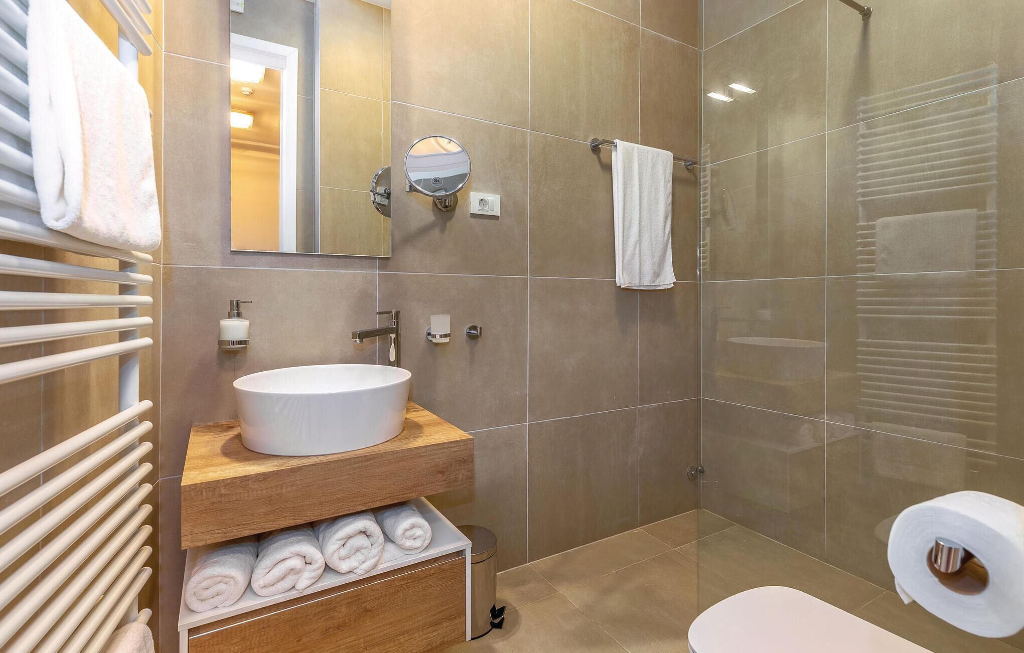 Un baño moderno con lavamanos blanco, tocador de madera, toallero y cabina de ducha con mampara de cristal.