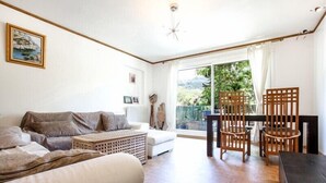 Een woonkamer met een bank, een bijzettafeltje en twee houten stoelen. Er is een balkon met uitzicht op groen.