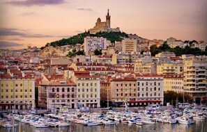 A coastal cityscape with a prominent hilltop church, dense urban buildings, and a marina filled with boats.