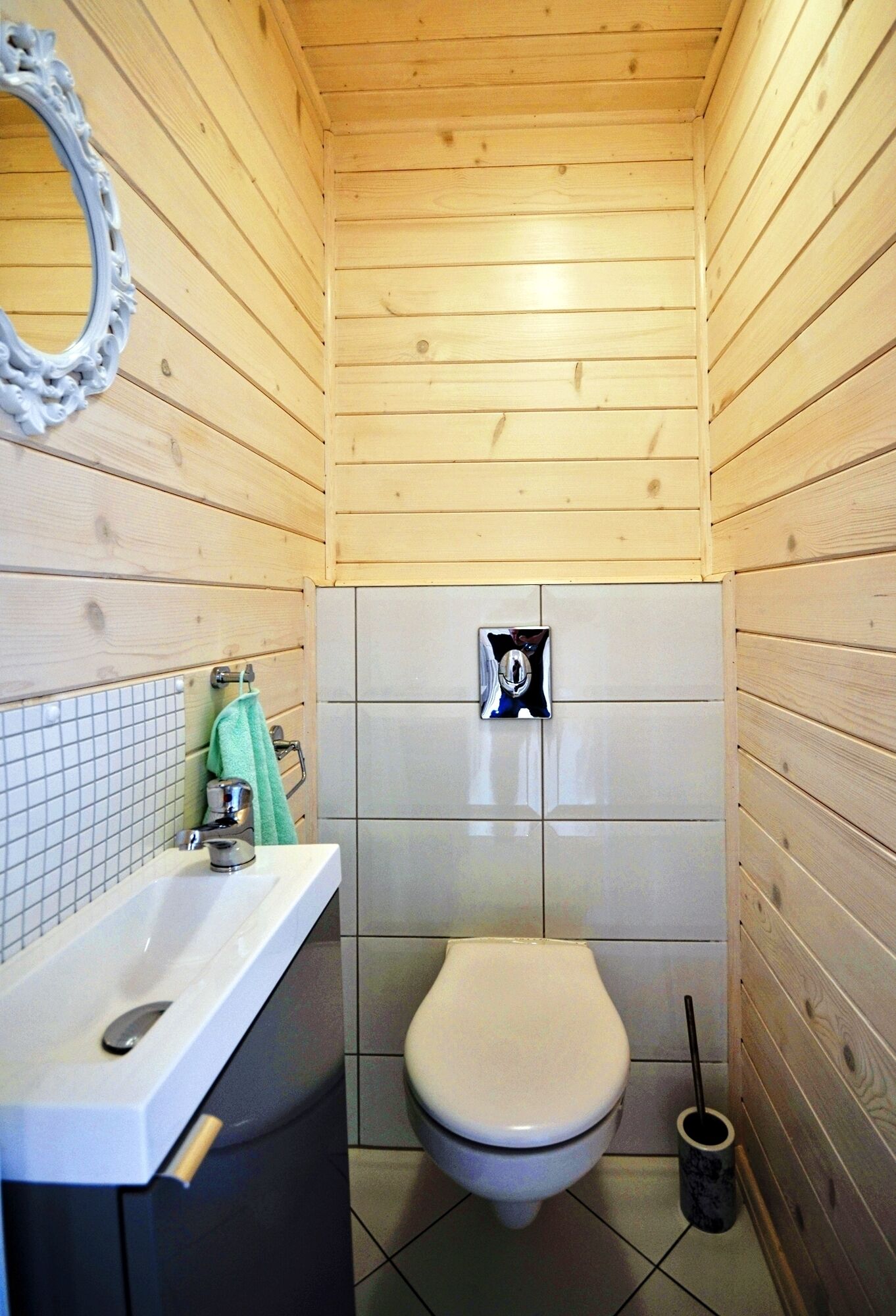 Een kleine, praktische badkamer met houten wanden, een wit toilet, een wastafel met spiegel en een betegelde vloer.