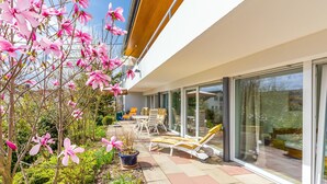 Uma casa moderna com um pátio com espreguiçadeiras e um jardim com flores cor-de-rosa.