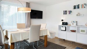Uma zona de refeições moderna com uma mesa posta para dois, uma televisão na parede e uma estante com livros e uma máquina de café.