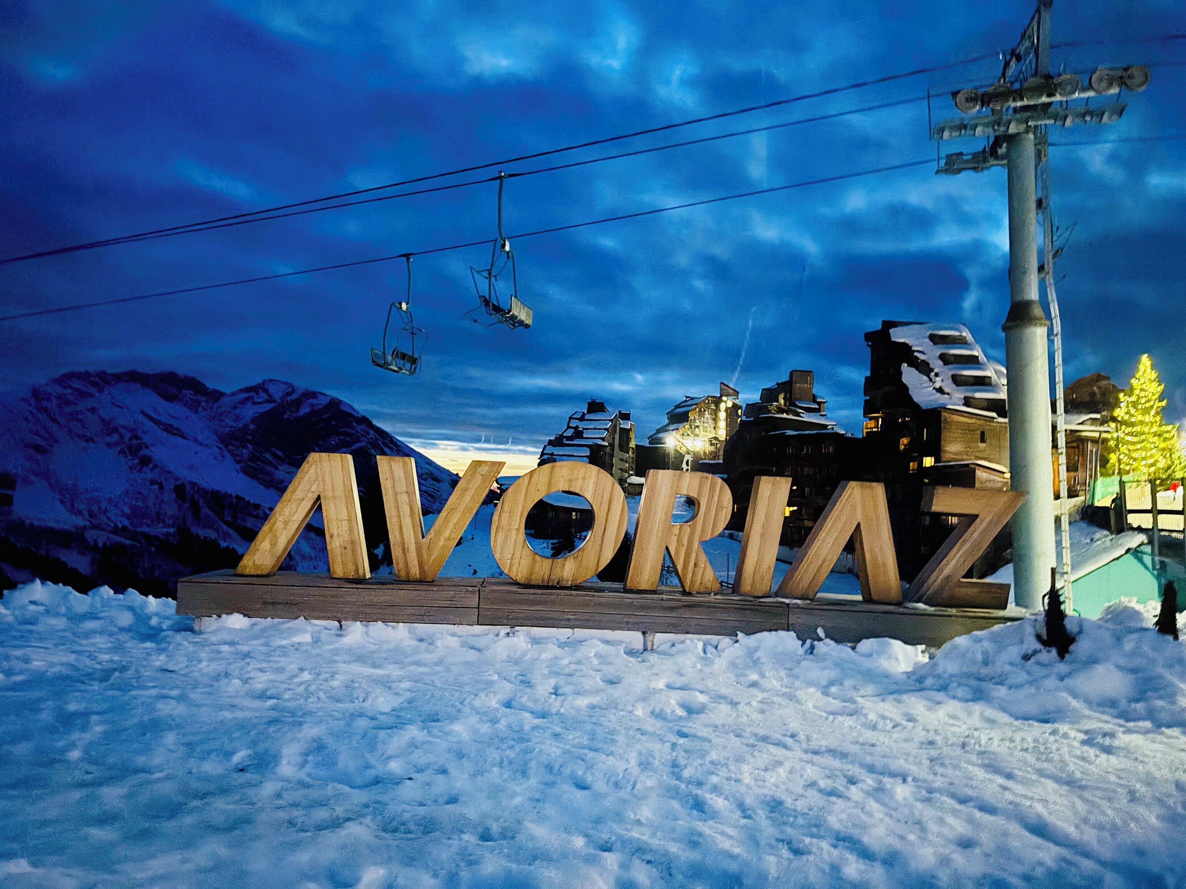 A wooden sign with the word "AVORIAZ" in large letters, set against a snowy landscape with mountains in the background and a ski lift in operation.