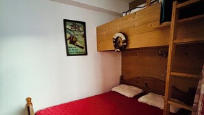 Una habitación pequeña con cama individual y colcha roja, cabecero de madera, una litera encima, escalera y un cartel en la pared.
