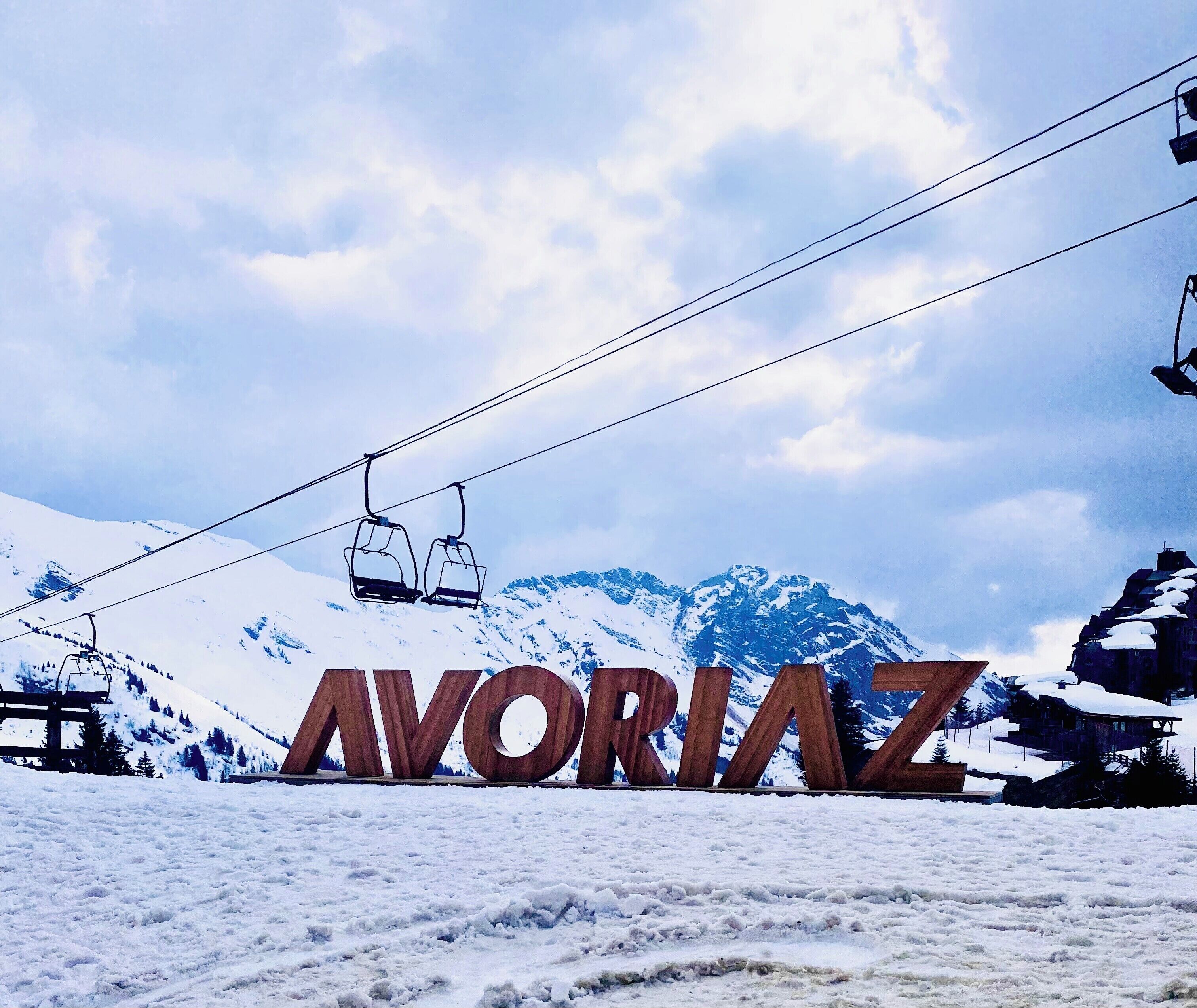 Une enseigne avec le mot « AVORAZ » écrit en grandes lettres, un télésiège en arrière-plan et des montagnes enneigées.