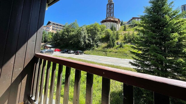 A view from a balcony overlooking a parking area with several cars, a church spire, and surrounding greenery. - Stylish Duplex with Mezzanine for 7, Steps from Slopes (Les Menuires)