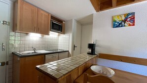 A compact kitchen with wooden cabinets, a microwave, a coffee maker, and a tiled backsplash. - Stylish Duplex with Mezzanine for 7, Steps from Slopes (Les Menuires)