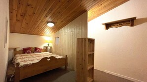 A bedroom with a wooden ceiling, a bed with a patterned bedspread, a nightstand with a lamp, and a wooden shelf unit. - Stylish Duplex with Mezzanine for 7, Steps from Slopes (Les Menuires)