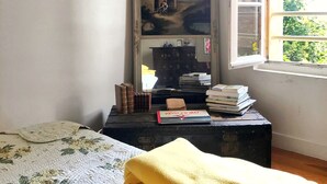 Una habitación con cama, ventana con vista a vegetación, espejo y una pequeña mesa con libros y un libro titulado 