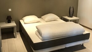 A modern bedroom with a large bed, a bedside table with a lamp, a small table with a decorative item, and a framed picture on the wall. - The clock Chalain 2 (Doucier)