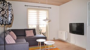 A living room with a grey sofa, two white side tables, a colorful striped rug, a black TV on the wall, and a window with blinds. - 1 bedroom cozy apartment in Varde (Varde)