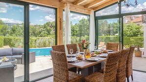 A dining area with wicker chairs, a table set for four, and a view of a swimming pool and greenery. - Amazing home in Kamenmost with sauna (Kamenmost)