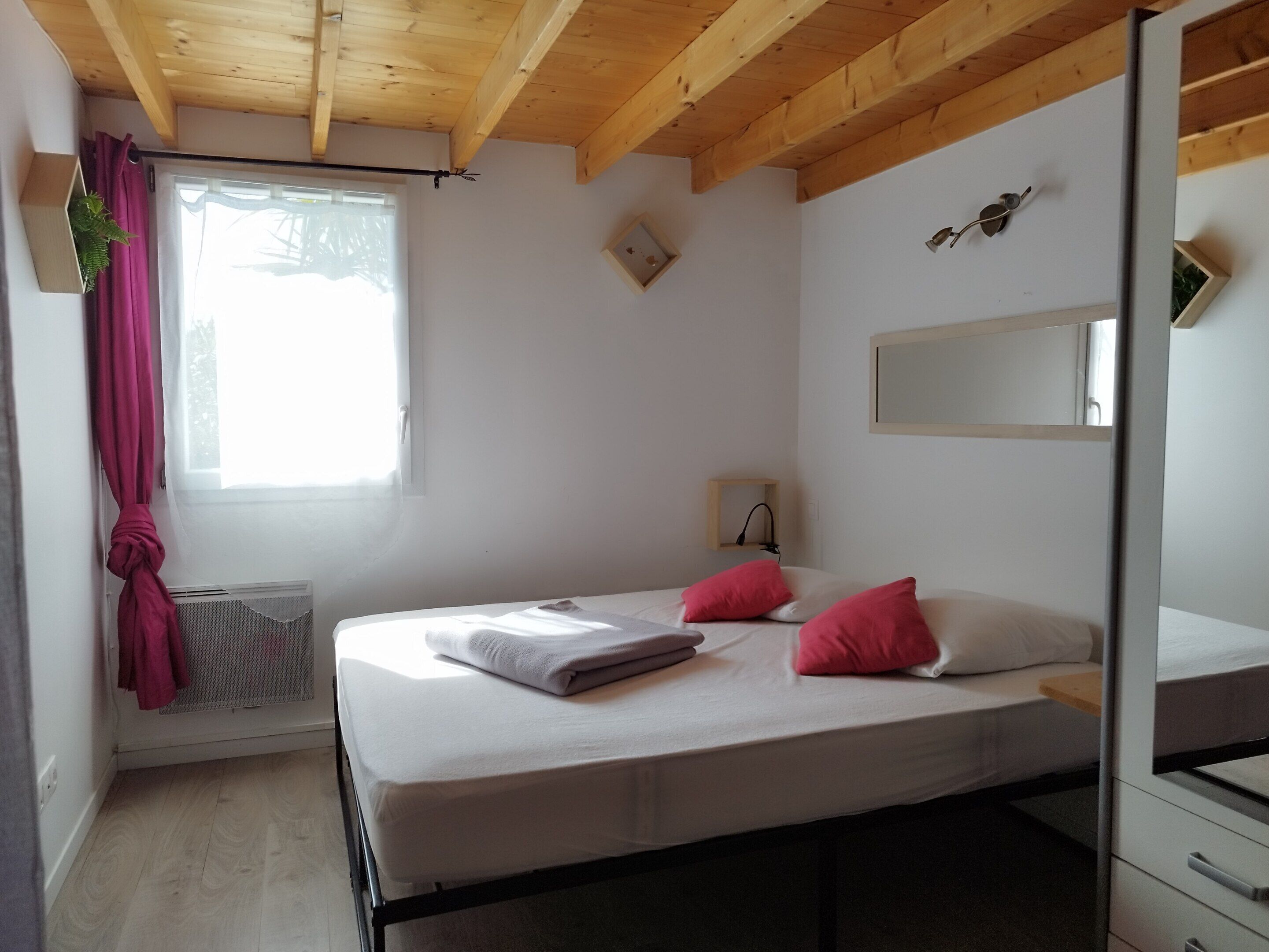 Une chambre à coucher avec un lit, une fenêtre avec des rideaux, un radiateur, un miroir et un plafond en bois.