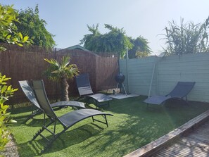 A backyard with artificial grass, two lounge chairs, a barbecue grill, a palm tree, and a wooden fence.