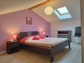 A bedroom with a sloped ceiling, a skylight, a bed with red pillows, bedside lamps, a small table with a scale, and a TV stand.