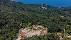 Oversigtsbillede af et resort med flere bygninger, swimmingpools og et skovområde omkring.