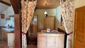 A room with wooden cabinets, a bed with floral curtains, a tiled floor, and a kitchen area in the background. - Farmhouse near the Baltic Sea (Upahl)