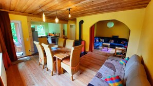 Un comedor con mesa y sillas de madera, una sala de estar con sofá y cojines coloridos, y una cocina al fondo.