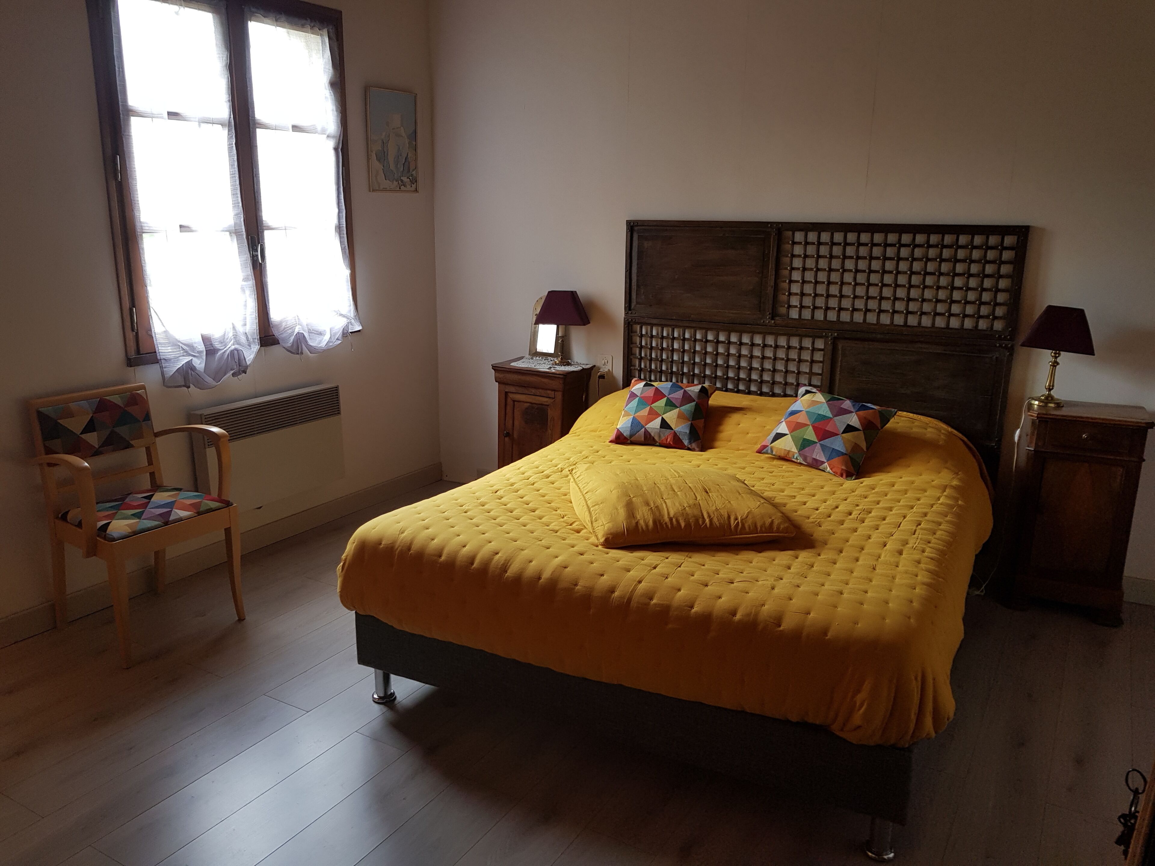 Un dormitori amb un llit gran, una cadira de fusta, una petita taula de nit i una finestra amb cortines.