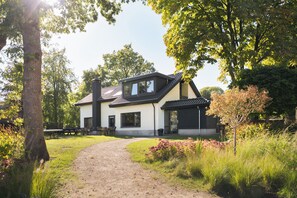 A two-story house with a dirt path leading to the entrance, surrounded by greenery and trees. - Group Villa for 24 people (Voorthuizen)