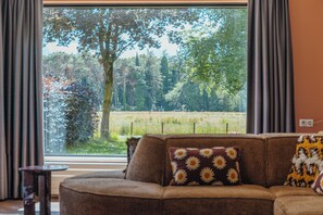 A living room with a large window, a brown sofa, and a small table. - Group Villa for 24 people (Voorthuizen)