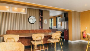 A dining area with a long table and chairs, a clock on the wall, and a kitchen in the background. - Group Villa for 24 people (Voorthuizen)