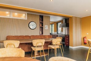 A dining area with a long table and chairs, a clock on the wall, and a kitchen in the background.