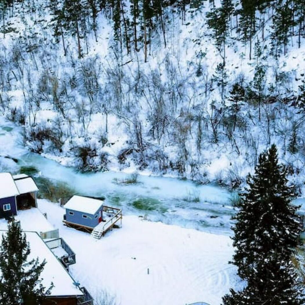 Eine Holzhütte in einem verschneiten Wald mit freiem Blick auf die umliegenden Bäume und einem kleinen Steg.