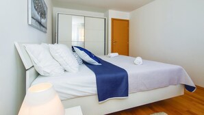 Cama bem arrumada com roupa de cama branca e azul, porta de madeira e luminária branca sobre piso de madeira.