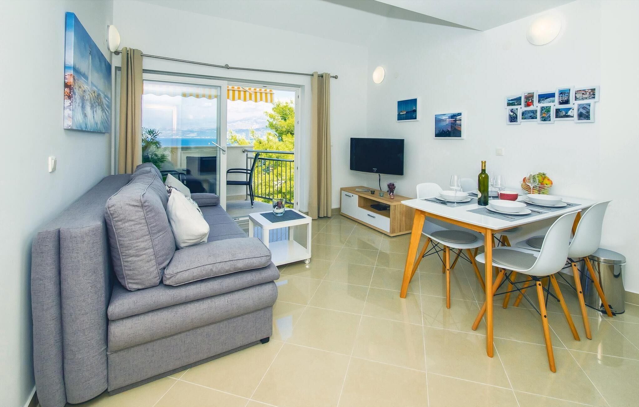 A modern living room with a grey sofa, a dining table with chairs, a TV, and a balcony with a view.