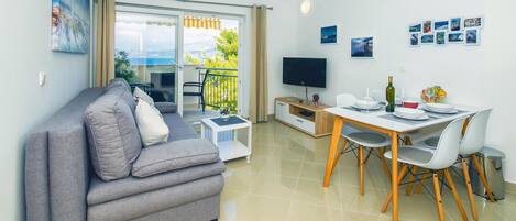 A modern living room with a grey sofa, a dining table with chairs, a TV, and a balcony with a view.