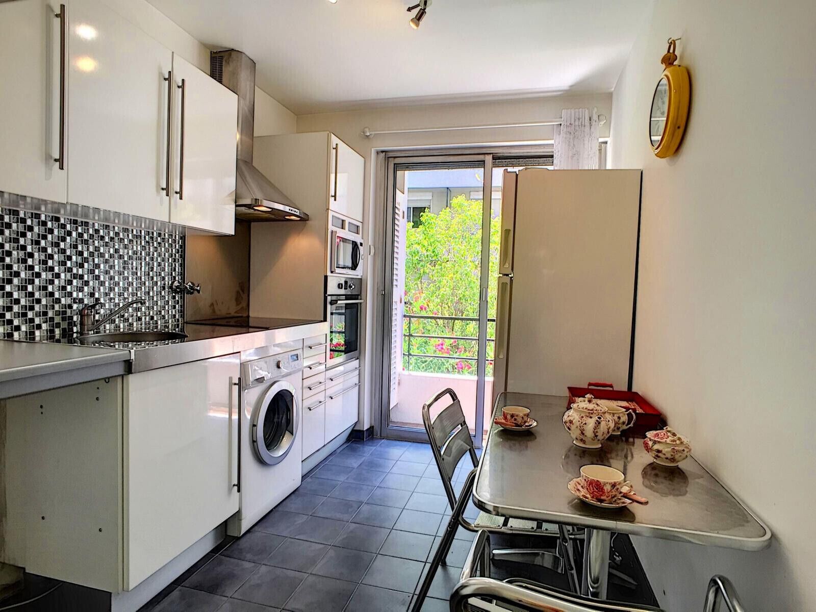 A compact kitchen with a washing machine, oven, and microwave. There's a dining table with tea cups.