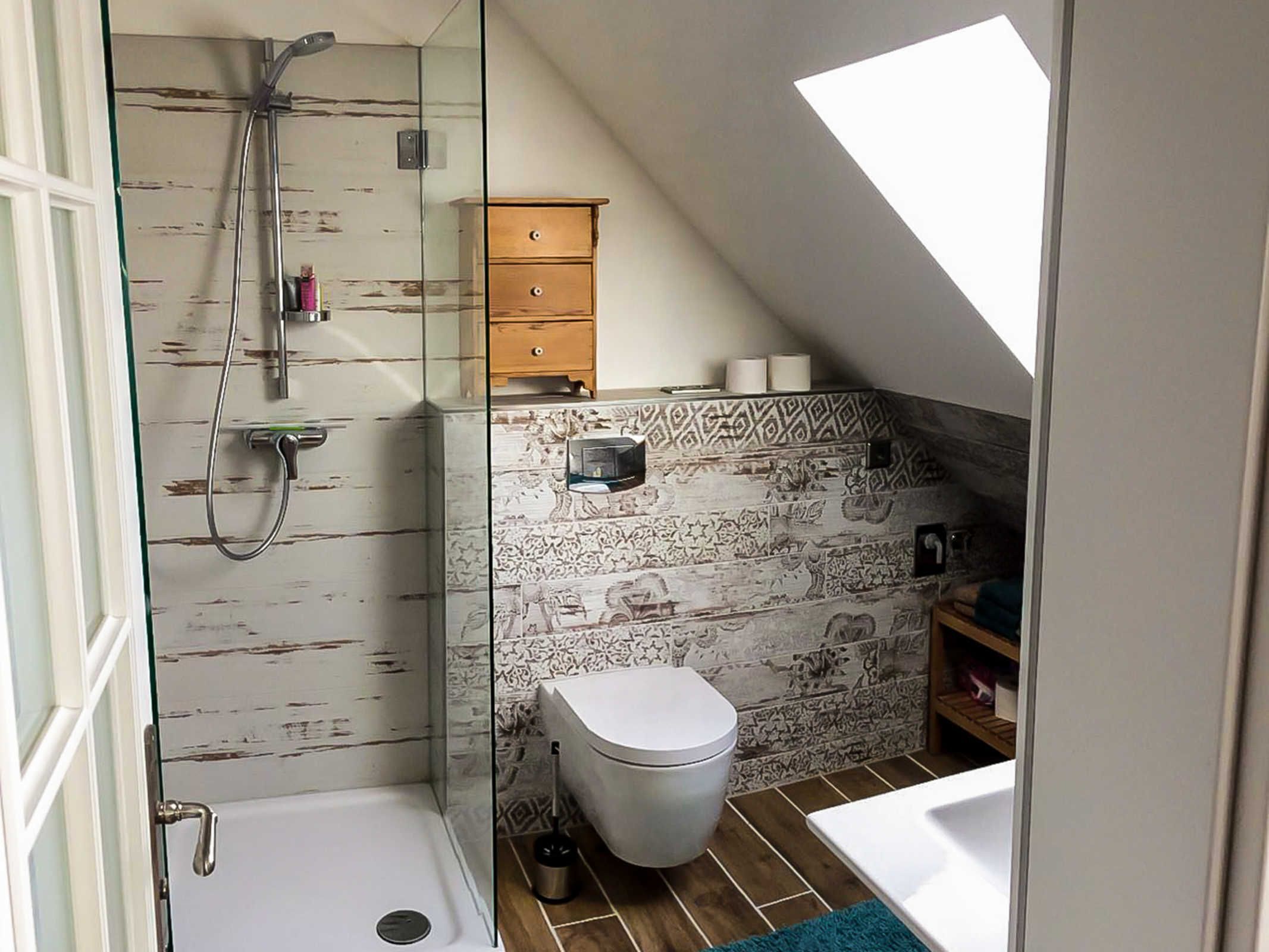 Ein kleines Badezimmer mit Dusche, Toilette und Waschbecken, mit schräg versiedeltem Dach und gemusterten Fliesen.